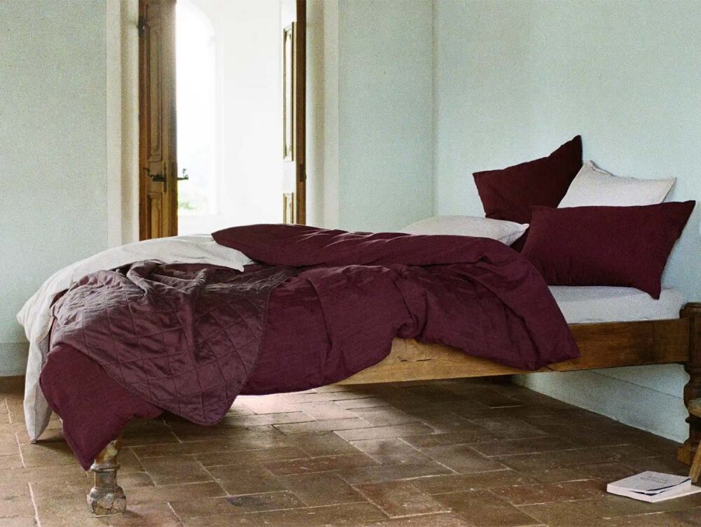 Ein ordentlich gemachtes Bett mit weinrotem Bettzeug und Kissen steht an einer hellen Wand in einem Raum mit Fliesenboden. Neben dem Bett liegt ein aufgeschlagenes Buch auf dem Boden, und durch eine offene Tür strömt Sonnenlicht.