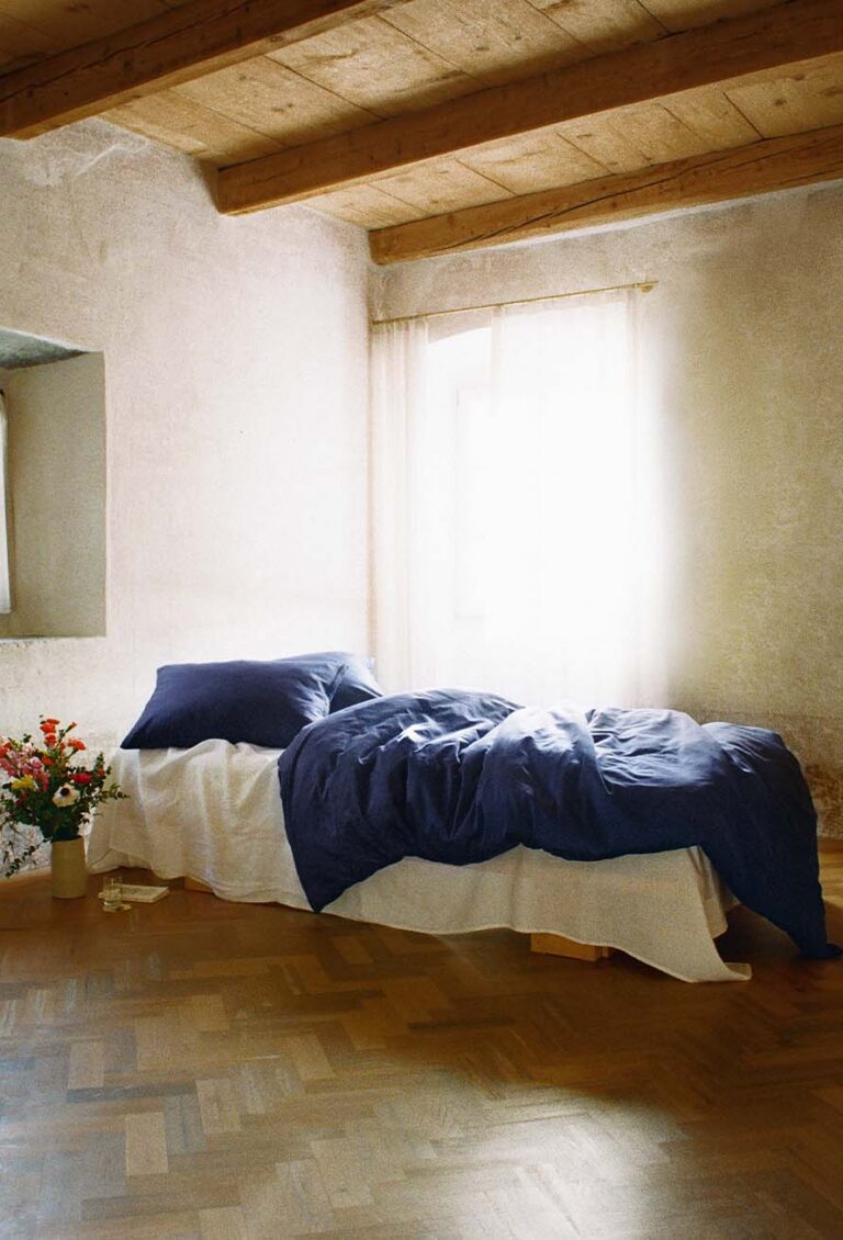 Ein sonnendurchflutetes Schlafzimmer mit einem Einzelbett mit weißem Laken, blauer Bettdecke und Kissen an einer strukturierten Wand mit Holzdecke. Ein Blumenstrauß in einer Vase steht auf dem Boden neben dem Bett.