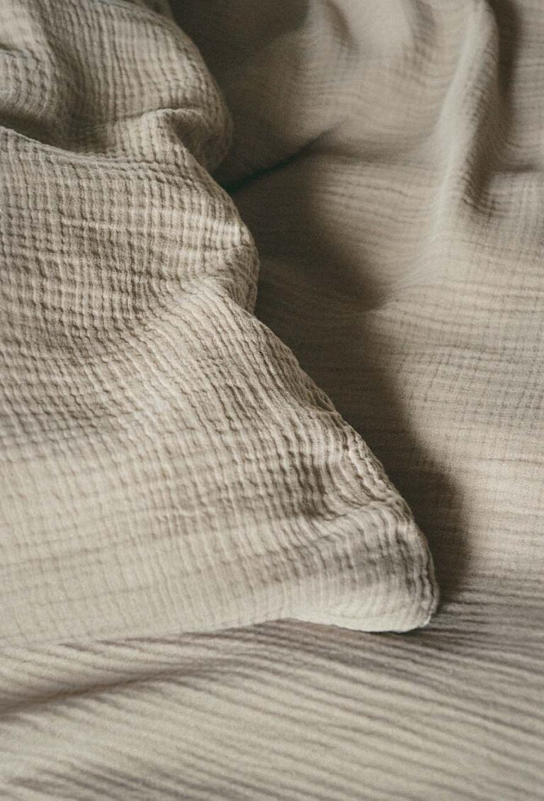 Nahaufnahme eines strukturierten, beigen Stoffes, möglicherweise eine Decke oder ein Bettüberwurf, mit weichen Falten und Wellen, die sanfte Schatten und ein gemütliches Aussehen erzeugen.