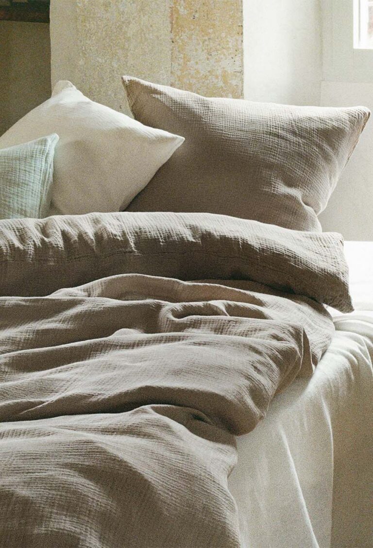 Ein ordentlich gemachtes Bett mit beigefarbener Bettwäsche und Kissen, vor einer rustikalen Wand und einem sonnendurchfluteten Fenster, schafft eine gemütliche und einladende Atmosphäre.