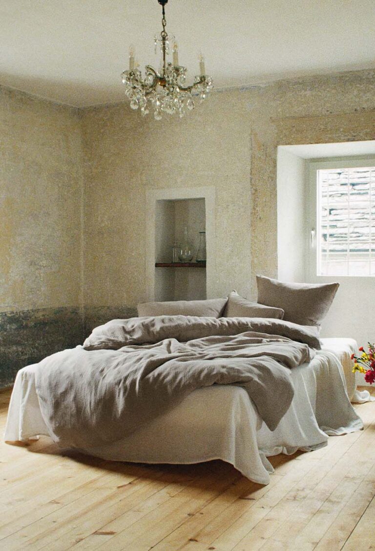 Ein minimalistisches Schlafzimmer mit einem zerknitterten Bett mit beiger Bettwäsche, einem Kristallkronleuchter über dem Bett, abgenutzten, strukturierten Wänden, einem Holzboden und einem Fenster, das natürliches Licht hereinlässt. Ein kleiner Blumenstrauß steht neben dem Bett.