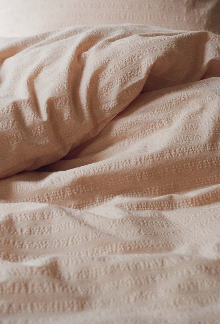 Eine Nahaufnahme von weicher, beiger Bettwäsche mit dezentem Strukturmuster, die sanfte Falten und Knicke im Stoff zeigt.
