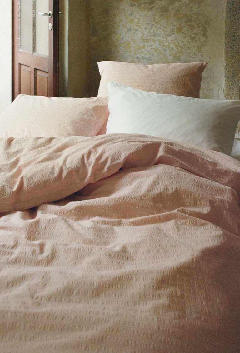 Ein gemütliches Bett mit einem blassrosa strukturierten Bettbezug, einem passenden Kissen und einem weißen Kissen, in einem sanft beleuchteten Raum mit einer rustikalen, verwitterten Wand und einer teilweise offenen Holztür im Hintergrund.