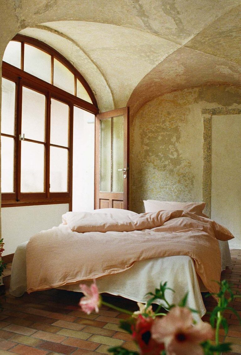 Ein gemütliches Schlafzimmer mit Gewölbedecken, großen Fenstern und einer Holztür. Das Bett ist mit hellrosafarbener Bettwäsche bezogen, und Sonnenlicht fällt ein und beleuchtet die rustikalen Wände und den gefliesten Boden. Die Blumen im Vordergrund sind unscharf.