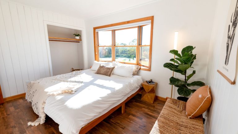 Helles Schlafzimmer mit einem großen Fenster, weißen Wänden und Holzböden. Ein Doppelbett mit weißem Bettzeug, eine Pflanze und ein offener Kleiderschrank schaffen einen ruhigen Rückzugsort - perfekt für Tipps und Tricks für ein kühles Schlafzimmer bei sonnigem Wetter.