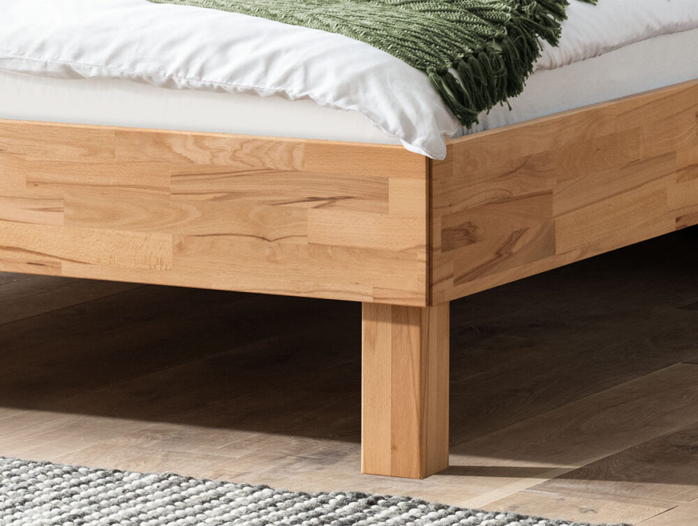 Eine Nahaufnahme eines hölzernen Bettgestells auf einem Hartholzboden mit einer weißen Matratze, weißem Bettzeug und einer grünen Decke. Im Vordergrund ist ein Teil eines grau-weißen Webteppichs zu sehen.