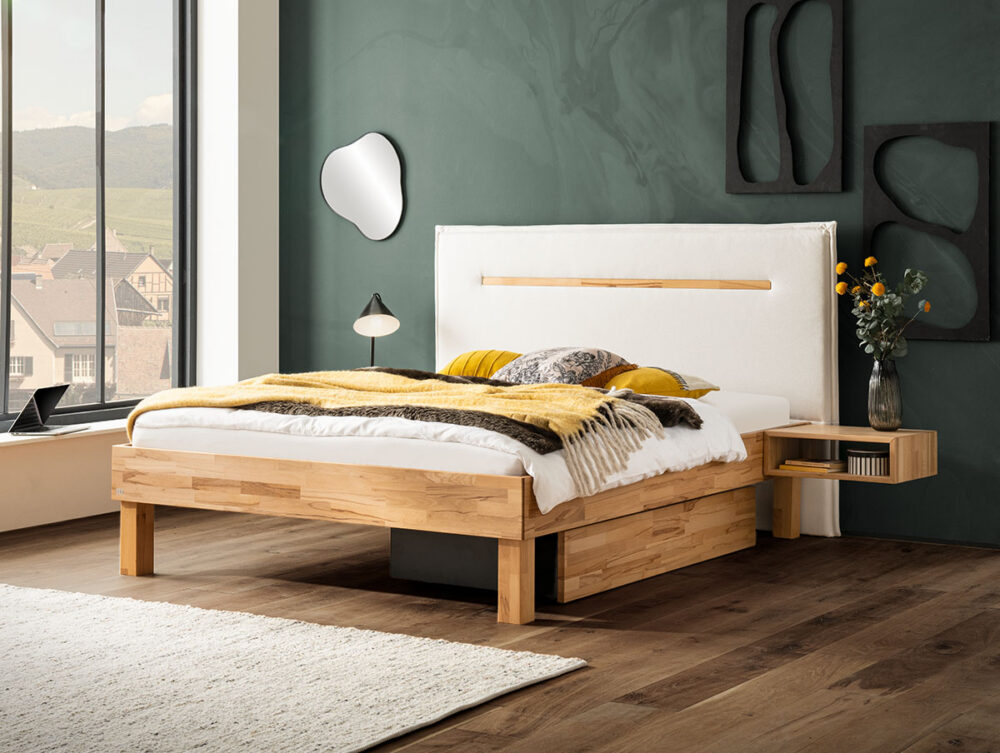 Modernes Schlafzimmer mit einem Holzbettgestell, weißer und gelber Bettwäsche, einem schwebenden Nachttisch mit Vase und Blumen, einer Lampe, Wandkunst und einem großen Fenster mit Blick auf Gebäude und Hügel. Grüne Akzentwand und Hartholzboden.