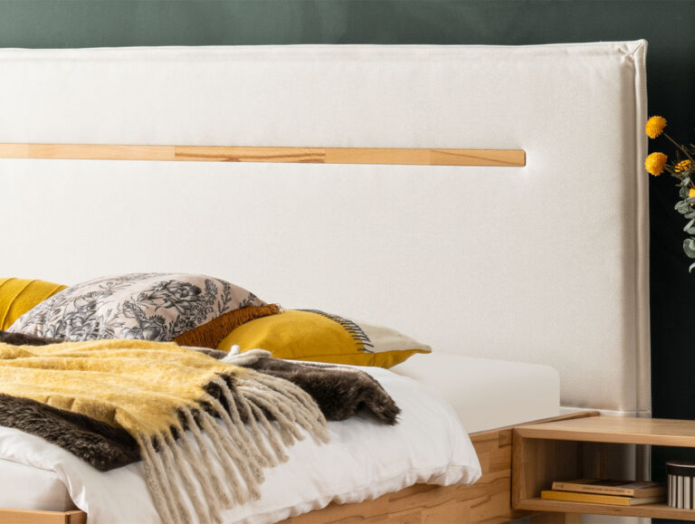 Ein modernes Bett mit einem hellen Holzrahmen und einem großen, gepolsterten weißen Kopfteil. Das Bett ist ordentlich mit weißer Bettwäsche, dekorativen gelben und grauen Kissen und einer gelben und grauen Fransendecke ausgestattet.