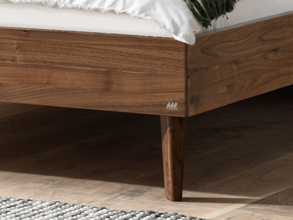 Nahaufnahme der Ecke eines hölzernen Bettrahmens mit spitz zulaufenden Beinen, weißem Bettzeug und einem Teil einer grünen Decke auf einem hellen Holzboden neben einem grau-weißen Teppich mit Struktur.