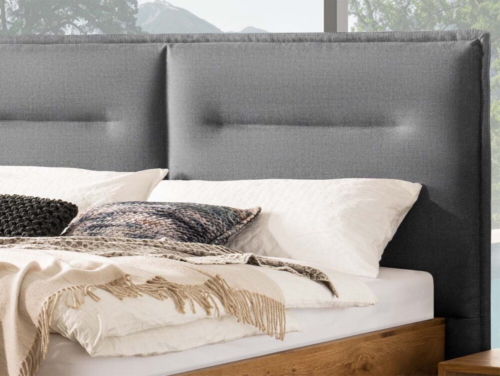 Ein ordentlich gemachtes Bett mit weißen und strukturierten Kissen, einer beigen Strickdecke und einem grauen, gepolsterten Kopfteil. Hinter dem Bett befinden sich große Fenster, die grüne Bäume und ferne Berge zeigen.