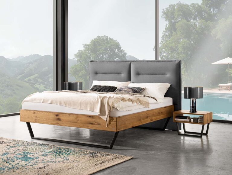 Ein modernes Schlafzimmer mit einem hölzernen Bettgestell, einem grau gepolsterten Kopfteil, weißer Bettwäsche, zwei Nachttischen mit Lampen, großen Fenstern mit Blick auf Bäume, Berge und einen Pool draußen.