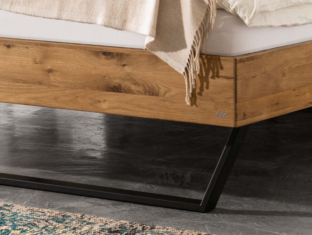 Nahaufnahme einer Bettecke mit einem Holzrahmen, schwarzen Metallbeinen und einer cremefarbenen Decke, die über den Rand drapiert ist, auf einem grauen Kachelboden neben einem gemusterten Vorleger.