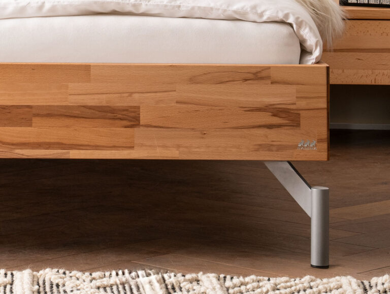 Nahaufnahme eines hölzernen Bettgestells mit hellbrauner Oberfläche und silbernen Metallfüßen. Auf dem Bett liegen eine weiße Matratze und Bettzeug, und auf dem Holzboden darunter ist ein Teil eines strukturierten Teppichs zu sehen.