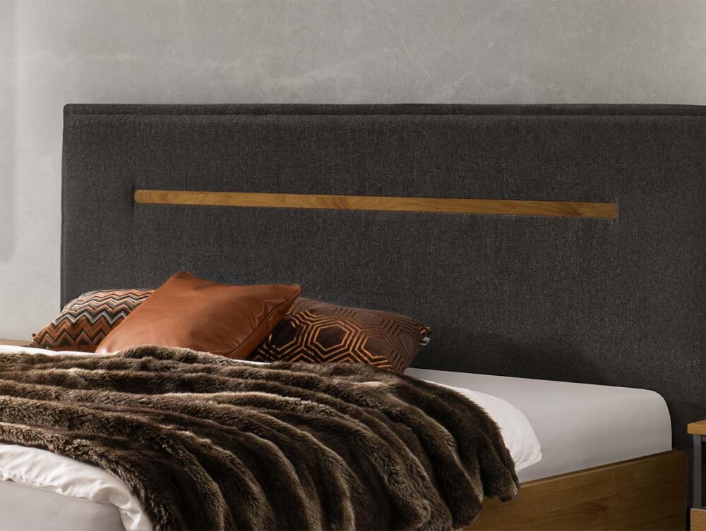 Ein modernes Bett mit einem hohen, dunkel gepolsterten Kopfteil mit Holzakzent, zwei dekorativen Kissen mit geometrischen Mustern und einer plüschigen braunen Kunstfelldecke, die über die weiße Bettwäsche drapiert ist.