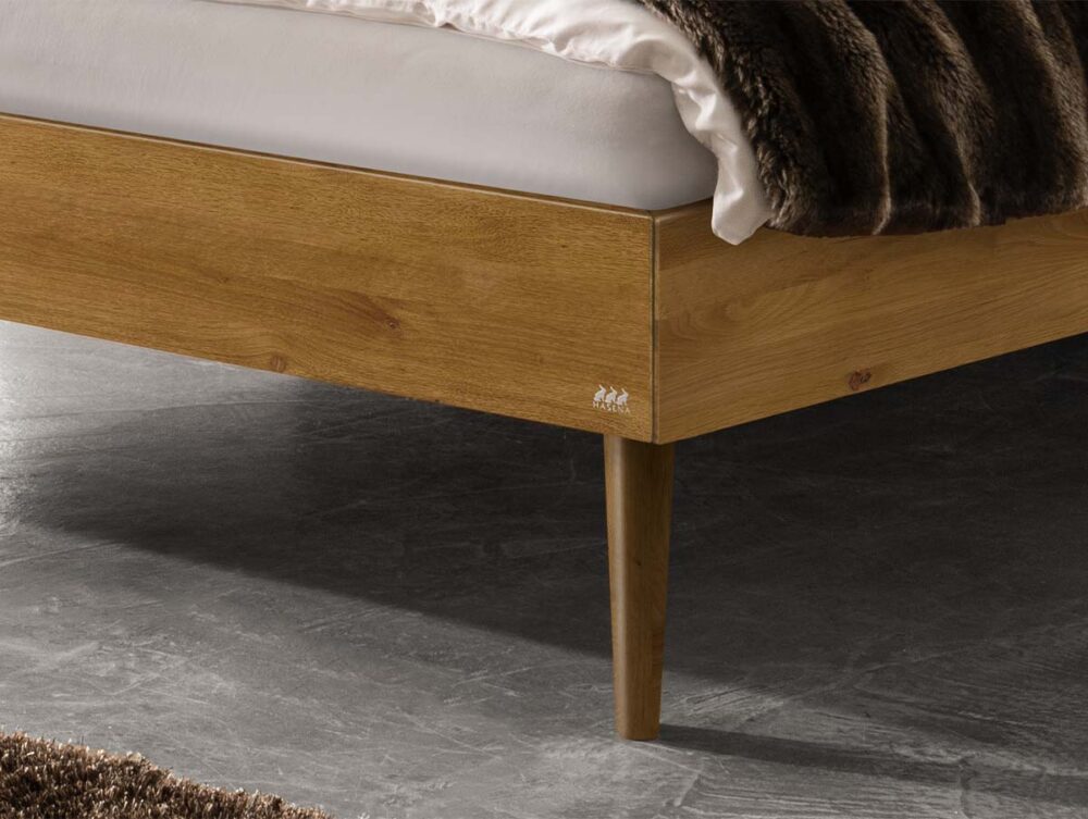 Nahaufnahme der Ecke eines hölzernen Bettrahmens mit spitz zulaufenden Beinen, einer weißen Matratze und einer über den Rand drapierten braunen Felldecke. Der Fußboden ist grau mit einer strukturierten Oberfläche, und ein brauner Teppich ist teilweise sichtbar.