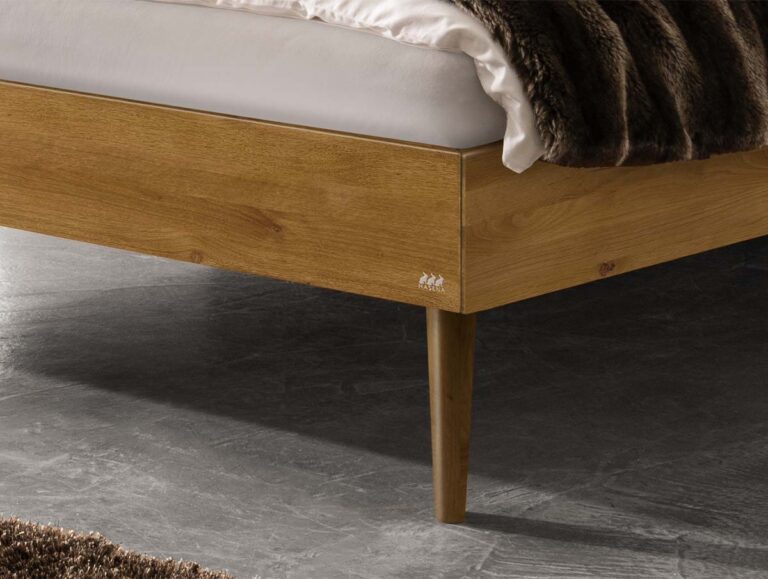 Nahaufnahme der Ecke eines hölzernen Bettrahmens mit spitz zulaufenden Beinen, einer weißen Matratze und einer über den Rand drapierten braunen Felldecke. Der Fußboden ist grau mit einer strukturierten Oberfläche, und ein brauner Teppich ist teilweise sichtbar.