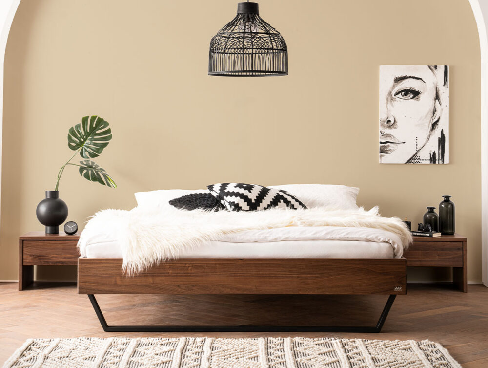 Ein modernes Schlafzimmer mit einem Holzbett, weißer Bettwäsche, einem schwarz-weißen Kissen, einer Pelzdecke, passenden Nachttischen, schwarzen Deko-Akzenten, einer Blattpflanze, abstrakter Wandkunst und einer schwarzen Pendellampe vor einer beigen Wand.