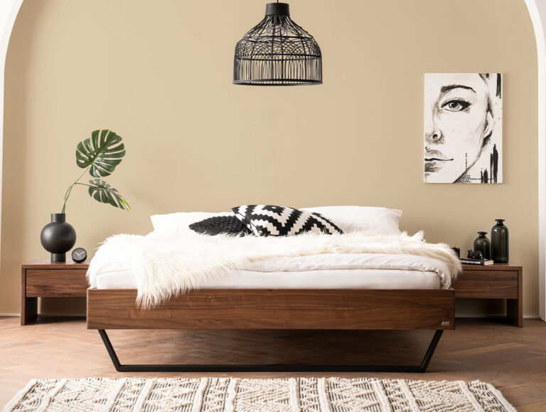 Ein modernes Schlafzimmer mit einem Holzbett, weißer Bettwäsche, einem schwarz-weißen Kissen, einer Pelzdecke, passenden Nachttischen, schwarzen Deko-Akzenten, einer Blattpflanze, abstrakter Wandkunst und einer schwarzen Pendellampe vor einer beigen Wand.