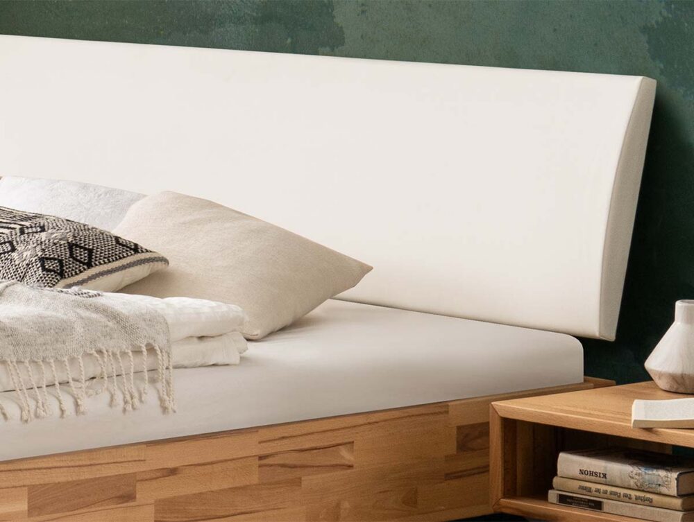 Nahaufnahme eines ordentlich gemachten Bettes mit weißem Kopfteil, hellem Bettzeug und Zierkissen. Auf einem hölzernen Nachttisch neben dem Bett stehen Bücher und eine kleine Keramikvase. Die Wand dahinter ist grün.