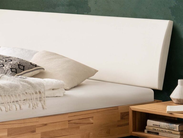 Nahaufnahme eines ordentlich gemachten Bettes mit weißem Kopfteil, hellem Bettzeug und Zierkissen. Auf einem hölzernen Nachttisch neben dem Bett stehen Bücher und eine kleine Keramikvase. Die Wand dahinter ist grün.