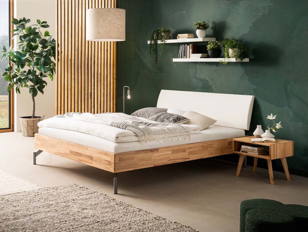 Ein modernes Schlafzimmer mit einem hölzernen Bettgestell, weißer Bettwäsche, einem hölzernen Nachttisch mit einer Lampe, einer grünen Akzentwand, Regalen mit Dekoration, einer großen Topfpflanze und natürlichem Licht aus einem Fenster.