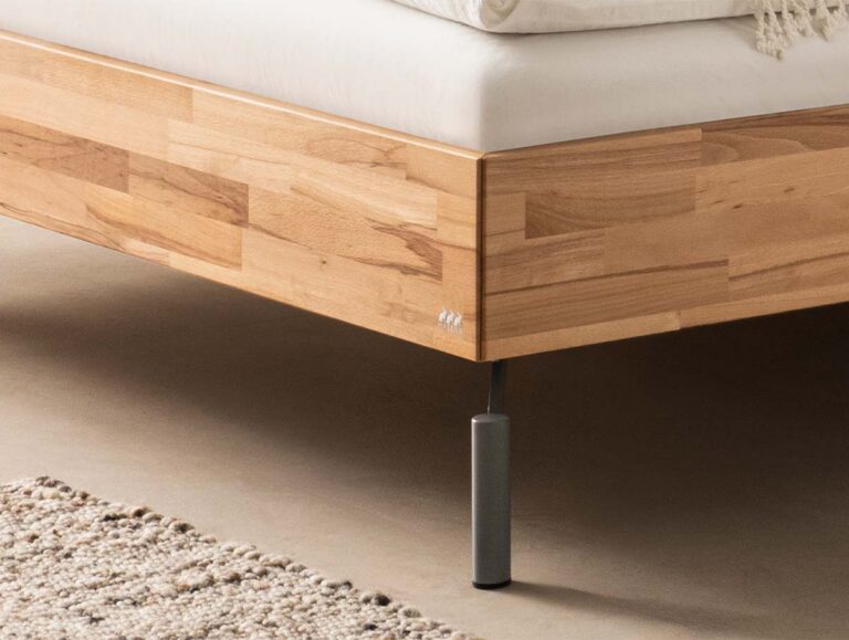 Nahaufnahme eines hölzernen Bettrahmens mit einer hellen Holzoberfläche, Metallfüßen und einer weißen Matratze. Auf dem Boden neben dem Bett ist ein Teil eines strukturierten beigen Teppichs zu sehen.