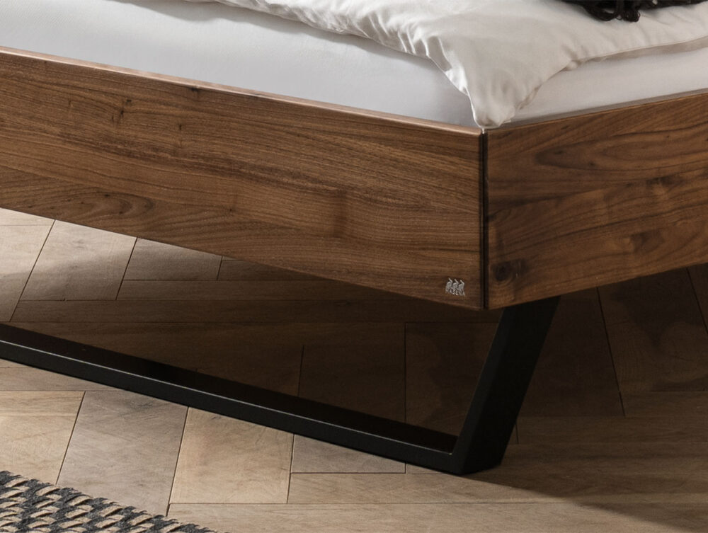 Nahaufnahme der Ecke eines hölzernen Bettgestells mit einer weißen Matratze. Der Rahmen hat ein modernes, kantiges Metallbein und steht auf einem hellen Holzboden mit Fischgrätmuster, auf dem ein Teil eines Teppichs zu sehen ist.