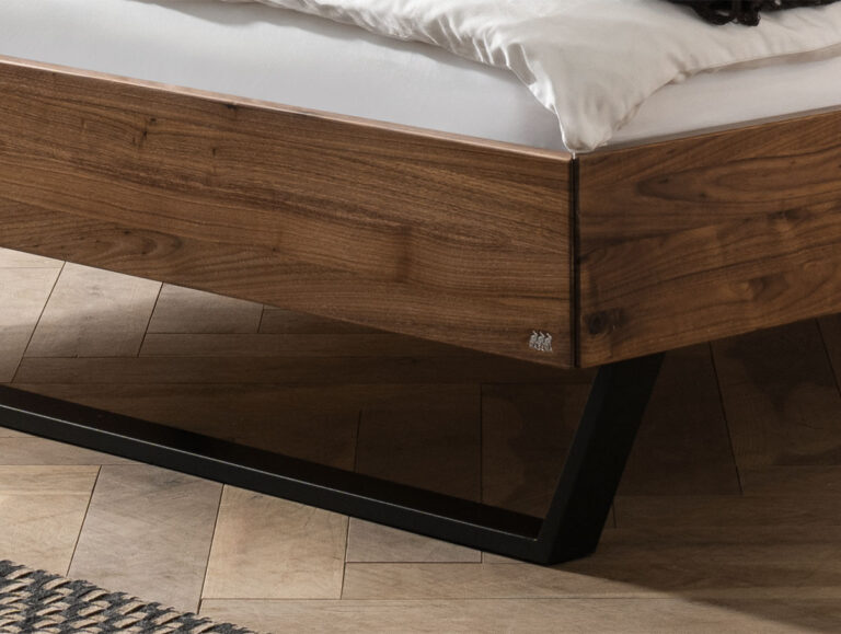 Nahaufnahme der Ecke eines hölzernen Bettgestells mit einer weißen Matratze. Der Rahmen hat ein modernes, kantiges Metallbein und steht auf einem hellen Holzboden mit Fischgrätmuster, auf dem ein Teil eines Teppichs zu sehen ist.