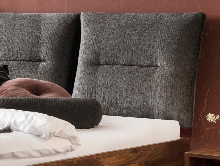 Nahaufnahme eines modernen Bettes mit einem dunkelgrauen Kopfteil aus strukturiertem Stoff, weißem Bettzeug und verschiedenen Kissen in Dunkelgrau und Staubrosa vor einer rötlich-braunen Wand. Ein kleines Stück eines hölzernen Nachttisches ist sichtbar.