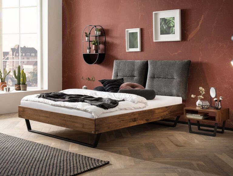 Ein modernes Schlafzimmer mit einem Holzbett mit grauem gepolstertem Kopfteil, weißer Bettwäsche und einer Überwurfdecke. Der Raum hat eine rotbraune Akzentwand mit gerahmter Kunst, Pflanzen, einen Nachttisch und ein großes Fenster, das natürliches Licht hereinlässt.
