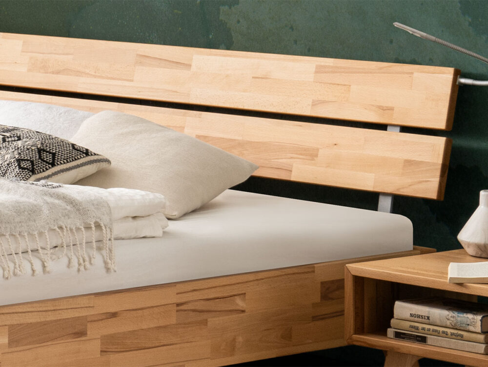 Ein modernes Holzbett mit Lattenrost, weißem Bettzeug, einer gefalteten Decke und einem gemusterten Kissen, daneben ein passender hölzerner Nachttisch mit Büchern und einem kleinen dekorativen Gegenstand.