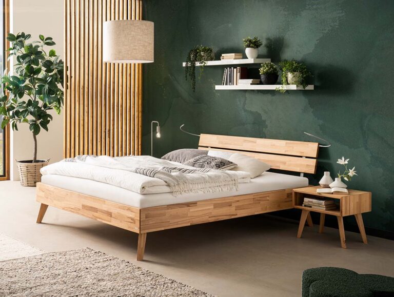 Ein modernes Schlafzimmer mit einem Holzbett, weißem Bettzeug und einem kleinen hölzernen Nachttisch. Die Wand ist dunkelgrün und hat zwei weiße Regale mit Pflanzen und Büchern. Eine hohe Pflanze steht neben einem Fenster mit Holzlatten.