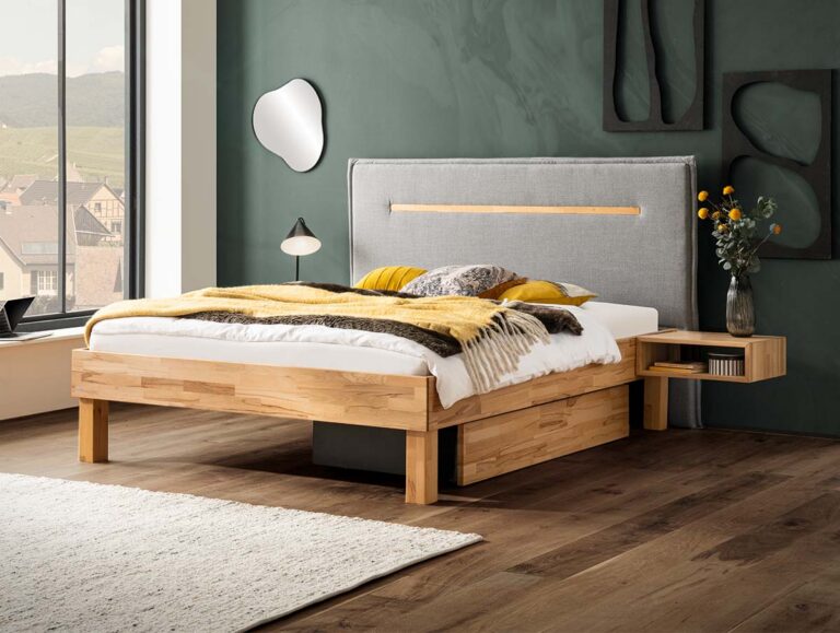 In einem modernen Schlafzimmer wird das Hasena Bett Bergamo "Anna" mit einem grau gepolsterten Kopfteil, einer senfgelben Decke und Kissen präsentiert. Der Look wird durch einen schwebenden Holznachttisch, frische Blumen, grüne Wände und große Fenster vervollständigt.