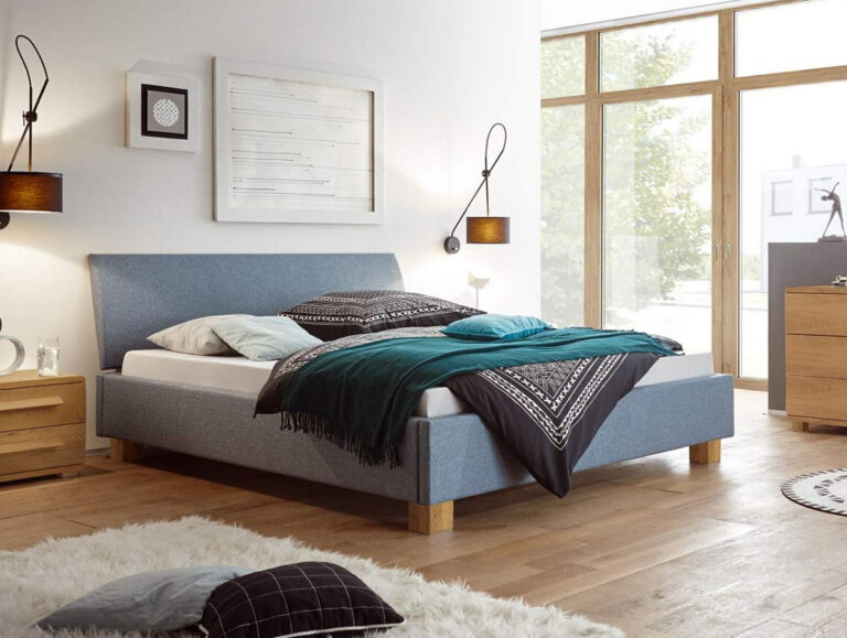 Ein modernes Schlafzimmer mit dem Hasena Bett "Dream-Line Lungo 28 Ivio 2", schwarz-weiß gemusterter Bettwäsche, tealfarbener Decke, hölzernen Nachttischen, Wandlampen, großen Fenstern und einem flauschigen weißen Teppich auf Holzfußboden.