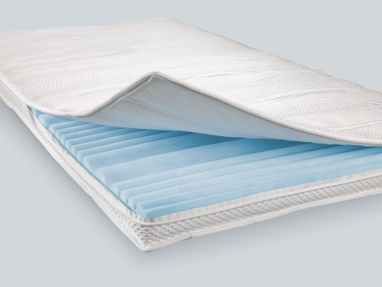 Nahaufnahme einer Matratze, bei der die oberste Schicht teilweise angehoben ist und eine hellblaue, strukturierte Schaumstoffschicht darunter zum Vorschein kommt, die den inneren Aufbau und die Materialien der Matratze veranschaulicht.
