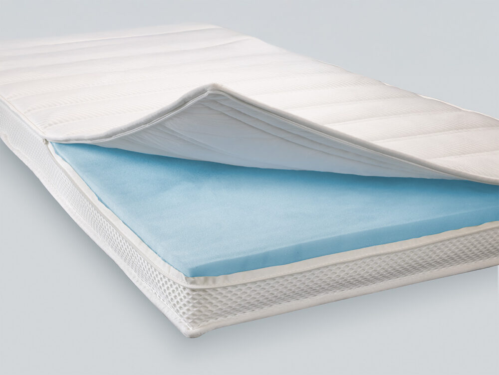 Nahaufnahme einer Matratze, deren weißer Steppbezug teilweise zurückgezogen wurde, so dass eine blaue Schaumstoffschicht im Inneren zum Vorschein kommt. Die Matratze steht vor einem schlichten hellen Hintergrund.