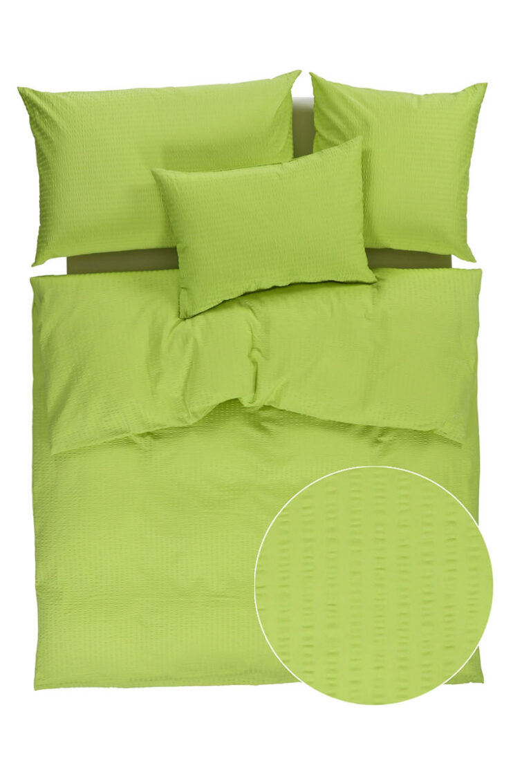 Ein ordentlich arrangiertes Bett mit einem grün strukturierten Bettbezug und drei passenden Kissen. Ein kreisförmiger Einsatz zeigt eine Nahaufnahme des subtilen Punktmusters der Stoffe.