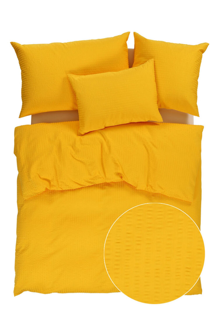 Ein hellgelbes Bettwäscheset mit einem strukturierten Muster, das eine Bettdecke, zwei Standardkissen und ein kleineres Kissen umfasst. Ein eingefügter Kreis zeigt eine Nahaufnahme des erhabenen Punktmusters des Stoffes.
