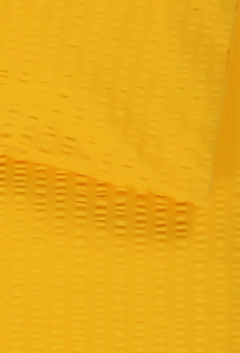 Nahaufnahme eines texturierten gelben Stoffes mit einem subtilen Gittermuster. Ein Stück Stoff überlappt ein anderes, wodurch eine diagonale Linie quer durch das Bild entsteht.