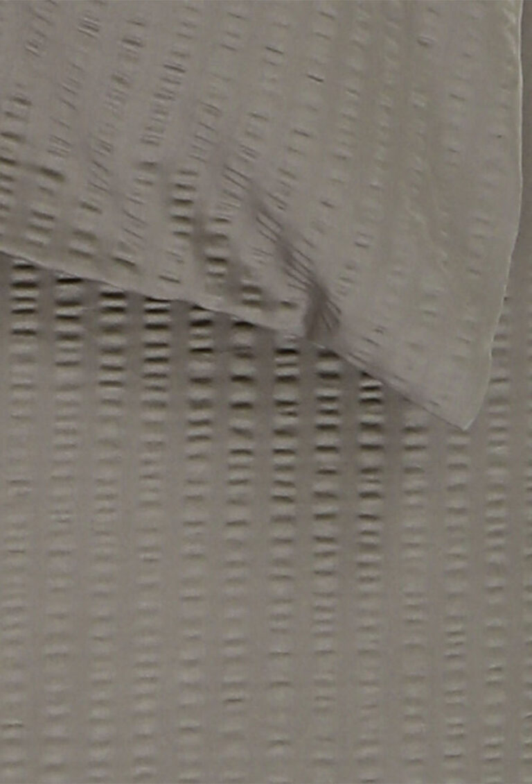 Eine Nahaufnahme eines hellgrauen, strukturierten Stoffes mit einem gewebten, gitterartigen Muster. Der Stoff scheint drapiert oder gefaltet zu sein, was weiche Schatten wirft und ein Gefühl von Tiefe erzeugt.