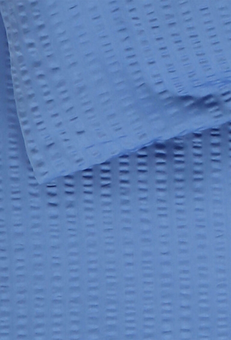 Nahaufnahme eines hellblauen strukturierten Stoffes mit einem gitterartigen, erhabenen Muster. Ein Stück Stoff ist über ein anderes gefaltet, wodurch ein leichter Schatten- und Schichteffekt entsteht.