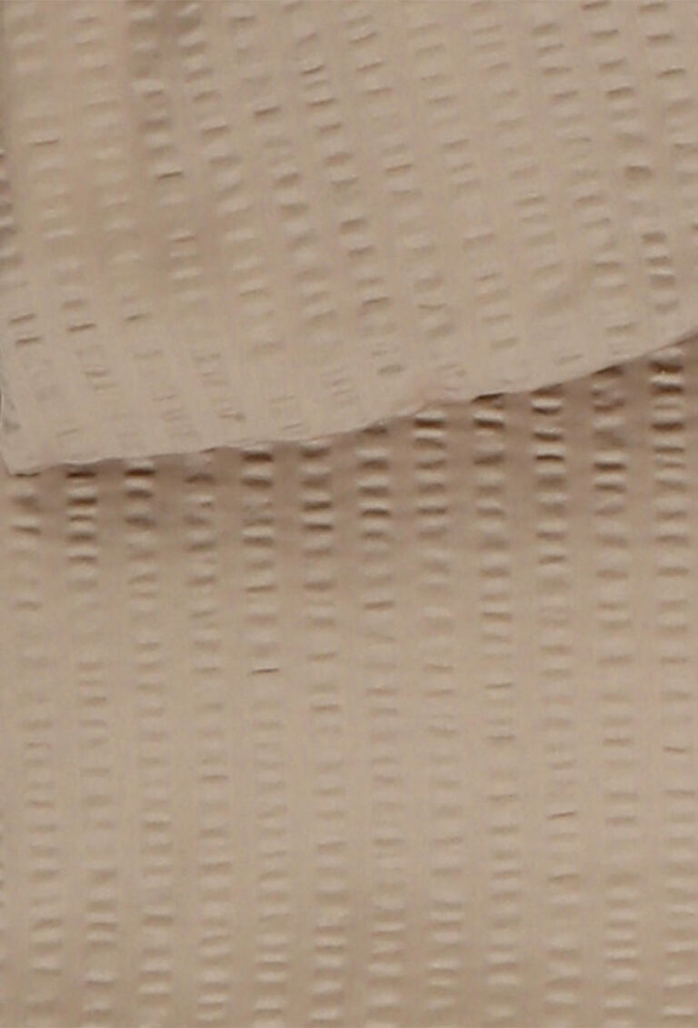 Nahaufnahme eines beigen Stoffes mit einem strukturierten, erhabenen, gepunkteten Streifenmuster, das vertikal und horizontal verläuft und ein gitterartiges Aussehen erzeugt. Der Stoff ist an der Oberseite leicht gefaltet.