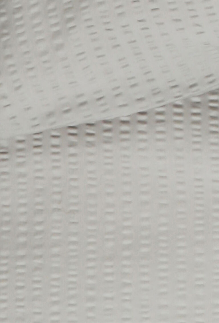 Nahaufnahme eines weißen, strukturierten Stoffes mit einem sich wiederholenden Muster aus kleinen, vertikalen, rechteckigen Vertiefungen. Der Stoff erscheint weich und im oberen Bereich des Bildes leicht gefaltet.