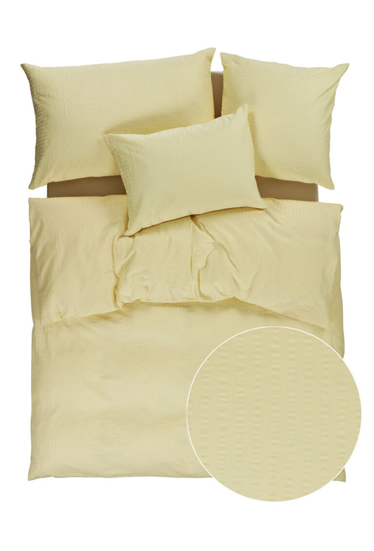 Ein ordentlich gemachtes Bett mit einem hellgelben, strukturierten Bettbezug und drei passenden Kissen; ein kreisförmiger Einsatz zeigt eine Nahaufnahme der subtilen, gepunkteten Textur des Stoffes.