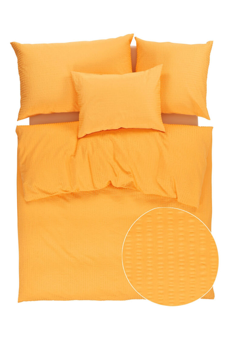 Ein ordentlich gemachtes Bett mit einem orangefarbenen strukturierten Bettbezug und drei passenden Kissen; ein kreisförmiger Einsatz zeigt eine Nahaufnahme des detaillierten Musters des Stoffes.