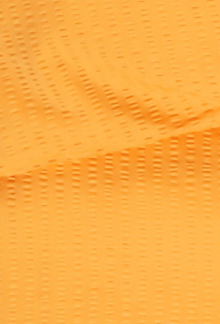 Nahaufnahme eines strukturierten orangefarbenen Stoffes mit kleinen, gleichmäßig verteilten vertikalen Rillen, die ein subtiles Muster auf der Oberfläche bilden. Der Stoff ist hell beleuchtet, so dass die Details des Gewebes und der Textur sichtbar werden.