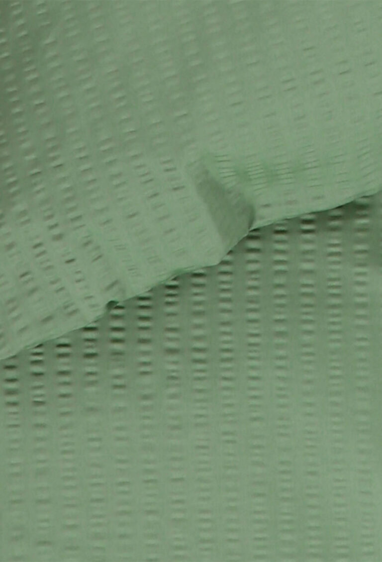 Nahaufnahme eines hellgrünen, strukturierten Stoffes mit einem subtilen Gitter- oder Schachbrettmuster, das Falten und Schatten zeigt, die die Oberfläche des Materials hervorheben.