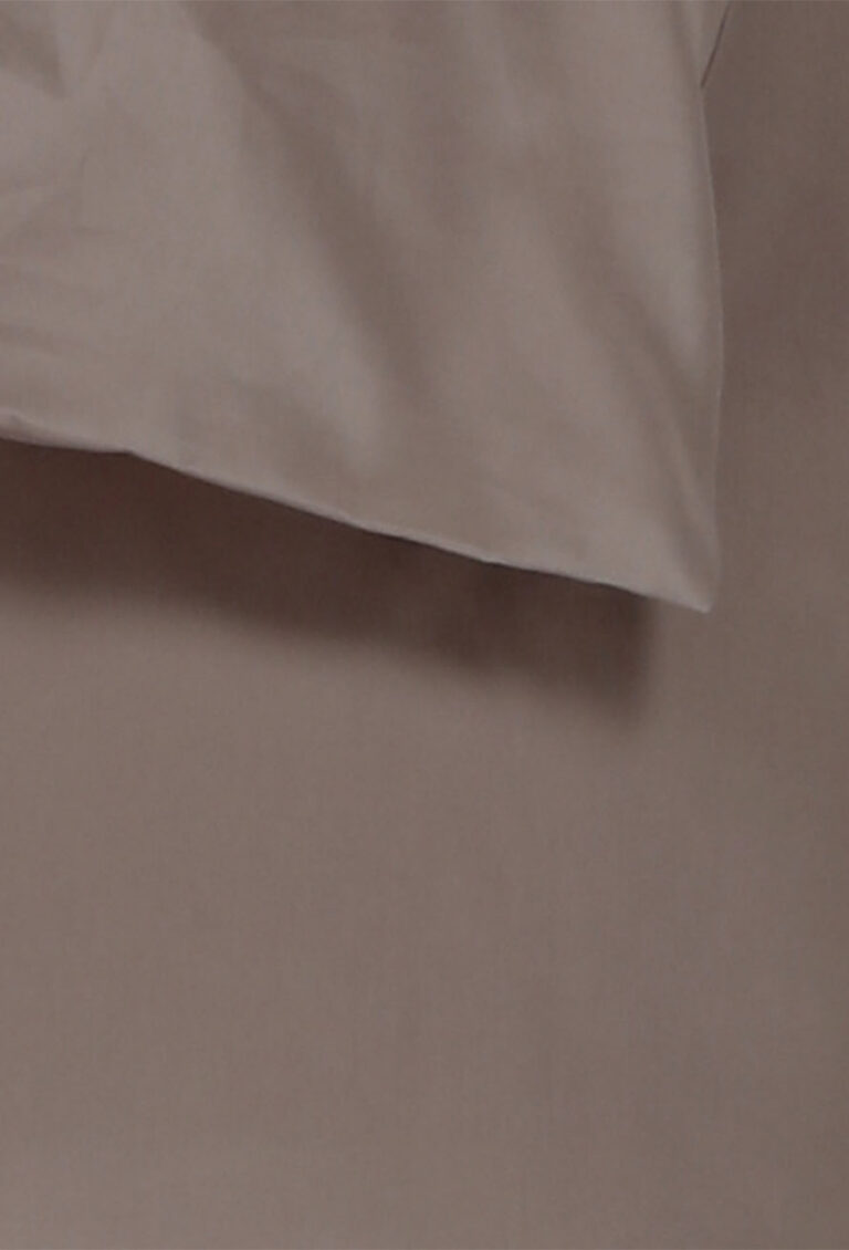 Eine Nahaufnahme eines hellbraunen oder taupefarbenen Stoffes, möglicherweise ein Bettlaken oder ein Kissenbezug, der über eine passende flache Oberfläche mit weichen Falten und Schatten drapiert ist.