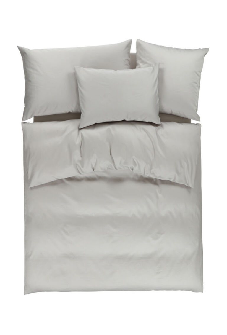 Ein ordentlich gemachtes Bett mit hellgrauer Bettwäsche, darunter eine Bettdecke, zwei große Kissen oben, ein Standardkissen rechts und ein Standardkissen in der Mitte, alles vor einem weißen Hintergrund.