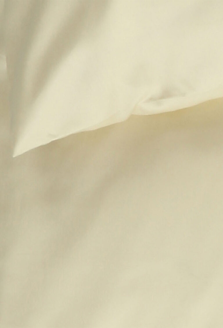 Eine Nahaufnahme eines cremefarbenen Kissens und eines dazu passenden Bettlakens, das einen glatten, weichen Stoff mit sanften Falten zeigt.